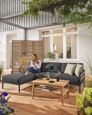 Eine Frau sitzt auf der Premium-Garten-Lounge-Ecke »Elin« mit einem flexiblen Paravent im Hintergrund. Eine schwarze LED-Akku-Outdoor-Steh- und Tischleuchte ist ebenfalls zu sehen, sowie ein Garten-Couchtisch.