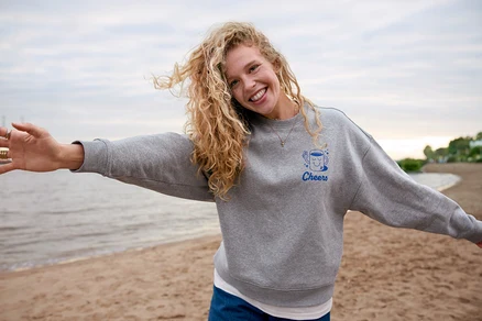 Frau mit lockigem Haar und grauem Pullover mit "Cheers"-Aufdruck steht am Strand und streckt die Arme aus.