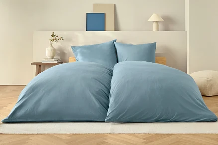 Blaue Bettdecke und zwei Kissen auf einem Bett auf einem weißen Teppich.