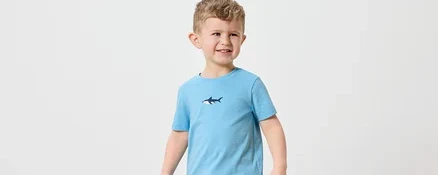 Ein kleiner Junge mit blonden Haaren trägt ein hellblaues T-Shirt mit einem Hai-Aufdruck und marineblaue Shorts. Er hält eine orangefarbene Gießkanne.