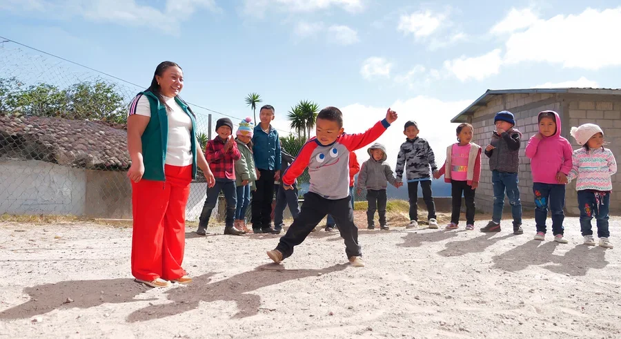 Eine Frau in grüner Weste und roter Hose lächelt einen Jungen in grau-rotem Shirt an, der auf einem Sandplatz tritt. Mehrere Kinder beobachten sie im Hintergrund unter blauem Himmel.