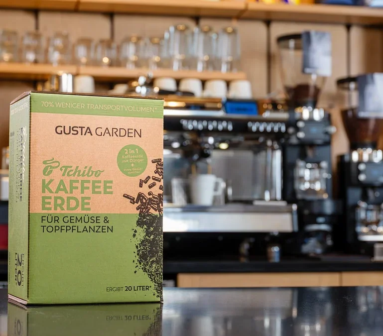 Eine Packung Tchibo Kaffee Erde steht vor einer Kaffeemaschine. Die Packung ist grün und braun und trägt die Aufschrift "Gusta Garden".