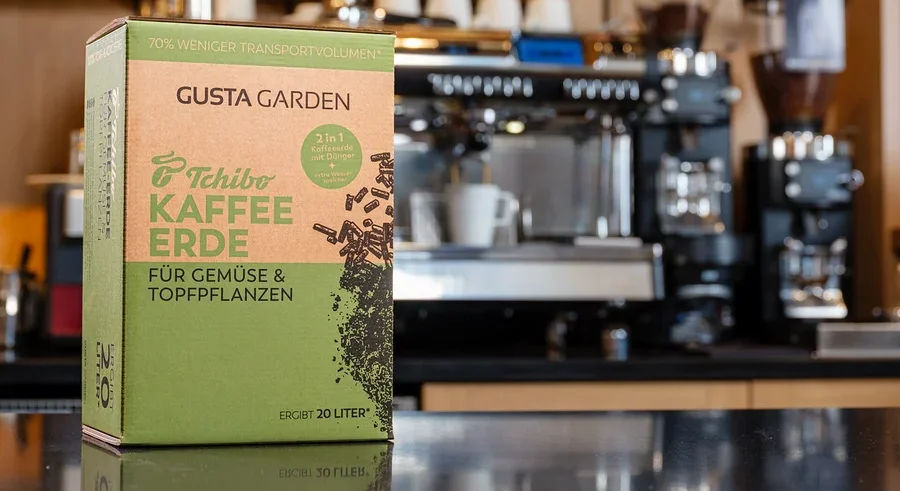 Eine Packung Tchibo Kaffee Erde steht vor einer Kaffeemaschine. Die Packung ist grün und braun und trägt die Aufschrift "Gusta Garden".