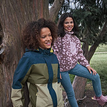 Zwei lächelnde Mädchen in einem Park, eines trägt eine mehrfarbige Jacke, das andere sitzt auf einem Baumast in einer gemusterten Fleecejacke und Kinder-Slim-Jeans ‒ Fit »Lotta«.