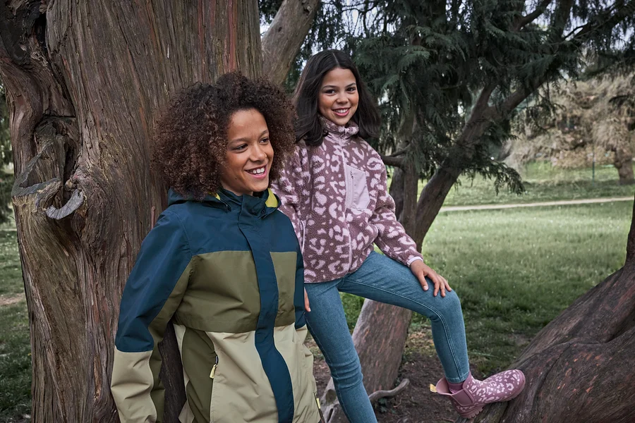 Zwei lächelnde Mädchen in einem Park, eines trägt eine mehrfarbige Jacke, das andere sitzt auf einem Baumast in einer gemusterten Fleecejacke und Kinder-Slim-Jeans ‒ Fit »Lotta«.