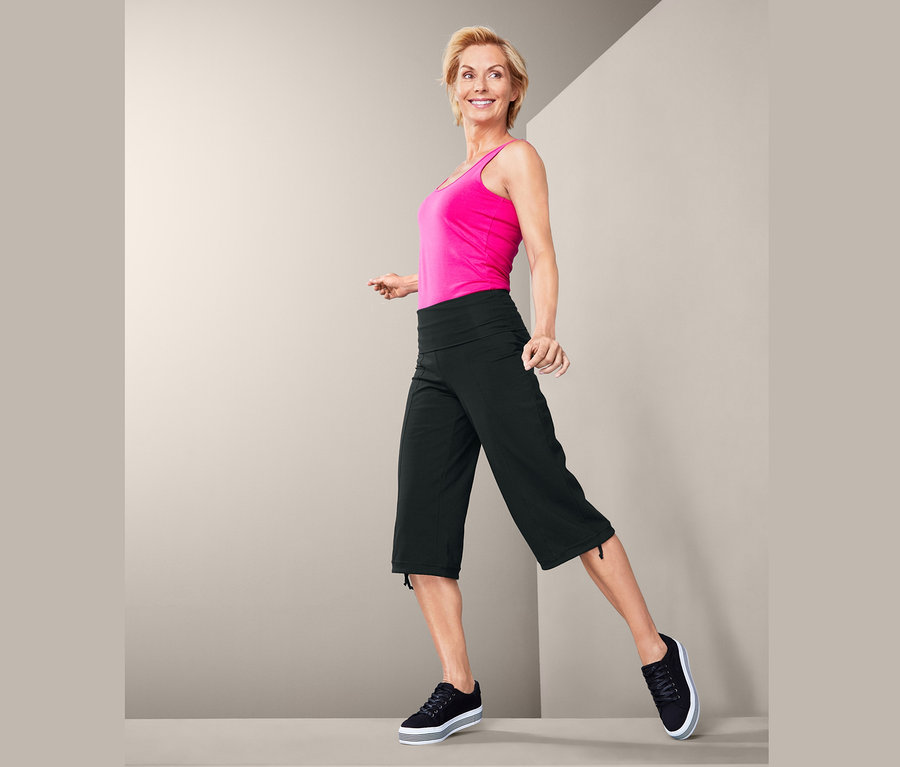 Frau in pinkem Top und 3/4-Sporthose steht vor grauem Hintergrund.