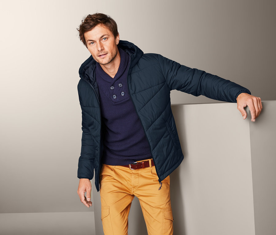 Mann, an eine Wand gelehnt, trägt eine Steppjacke, einen Pullover und gelbe Hosen.
