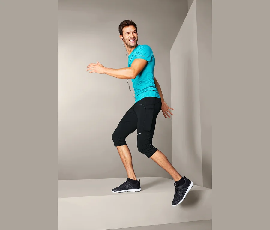 Lächelnder Mann in türkisfarbenem T-Shirt und 3/4-Lauftight posiert vor grauem Hintergrund.