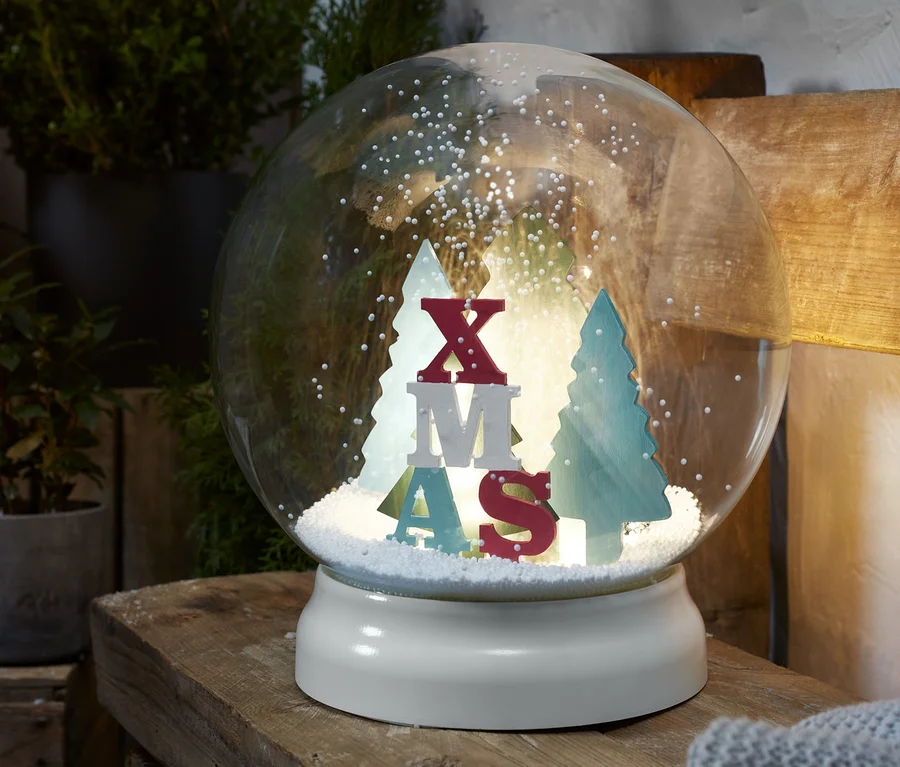 Eine LED-Outdoor-Schneekugel mit der Aufschrift XMAS und Bäumen im Inneren, die auf einer Holzoberfläche steht.