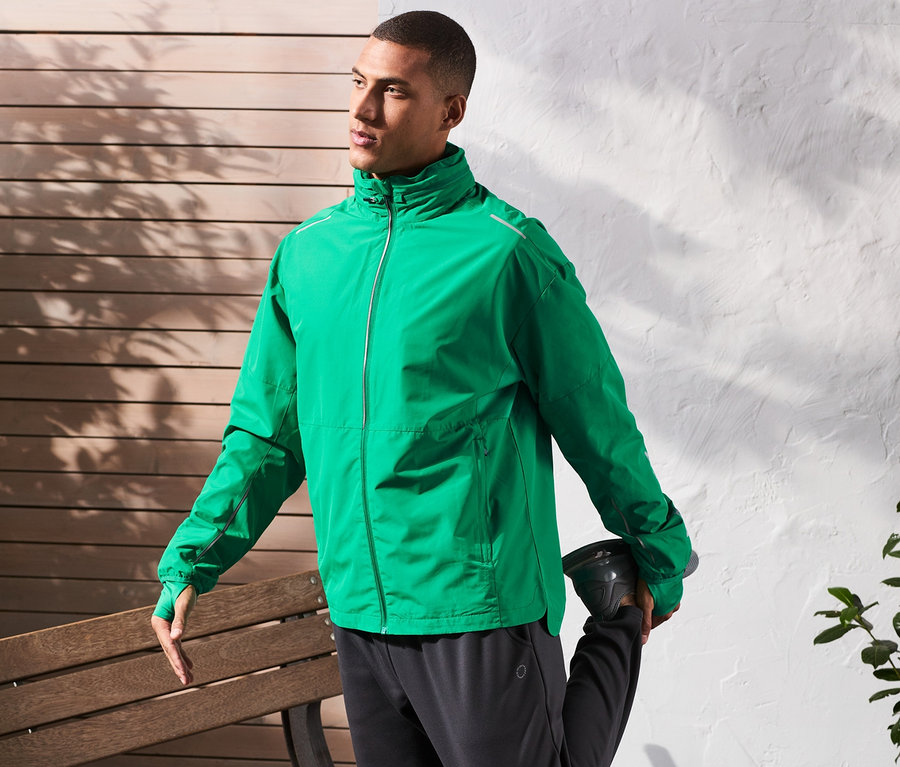 Mann dehnt sein Bein an einer Bank, trägt eine Windprotection-Funktionsjacke.