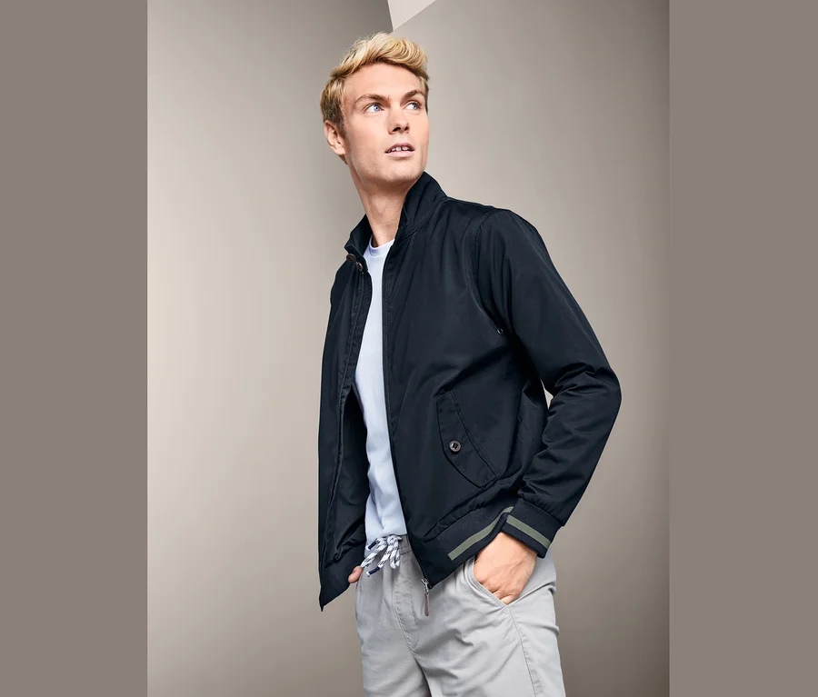 Ein Mann mit blonden Haaren trägt eine blaue Blouson und ein hellblaues T-Shirt.