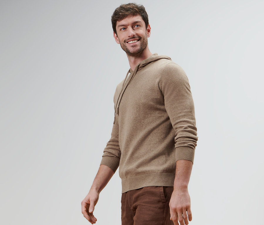 Ein lächelnder Mann trägt einen Strick-Hoodie und braune Hosen.