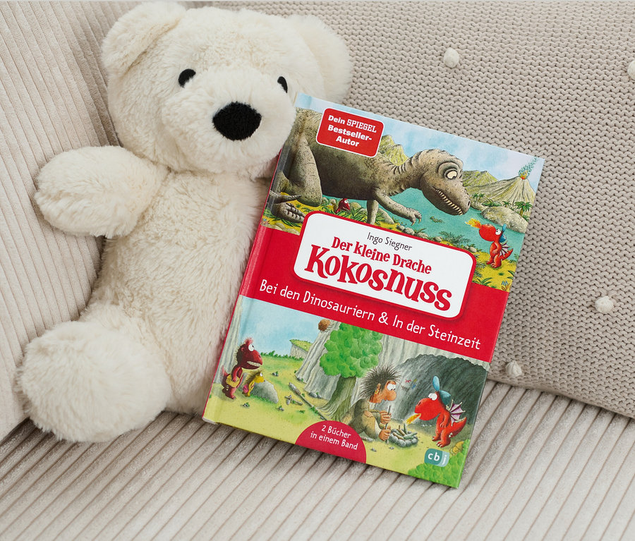 Ein Teddybär liegt neben dem Buch "Der kleine Drache Kokosnuss ‒ bei den Dinosauriern & In der Steinzeit".