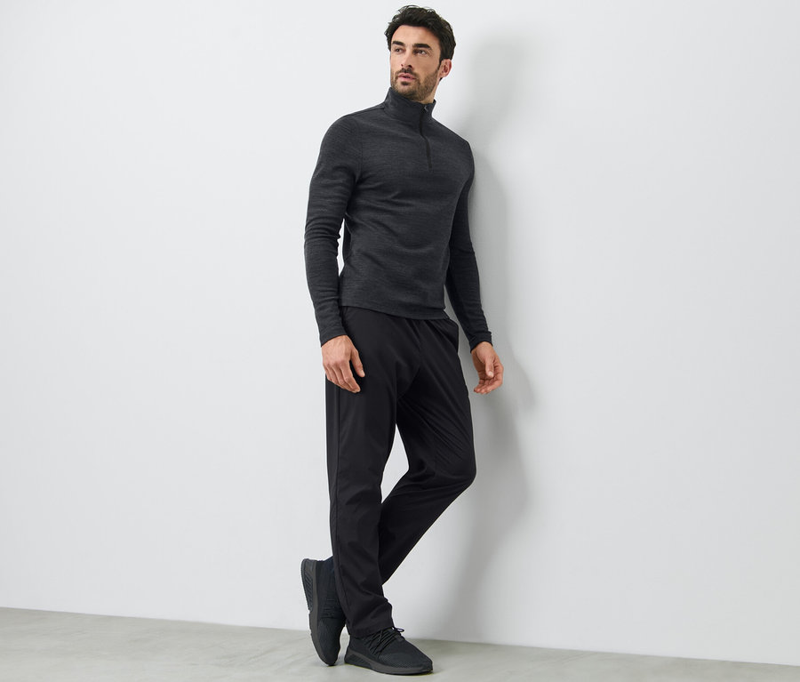 Mann lehnt an einer Wand und trägt eine schwarze Funktionshose und ein langärmeliges Oberteil.