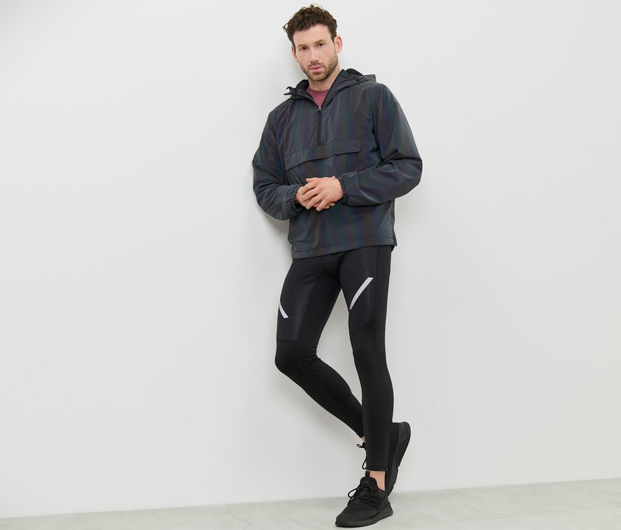 Mann lehnt an Wand, trägt Windbreaker Rainbow Reflective, schwarze Leggings und Turnschuhe.