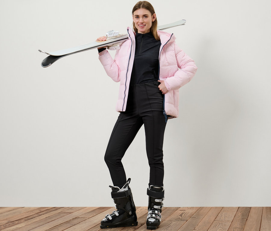 Frau posiert in Softshell-Ski-Tights und Skischuhen mit Skiern über der Schulter.