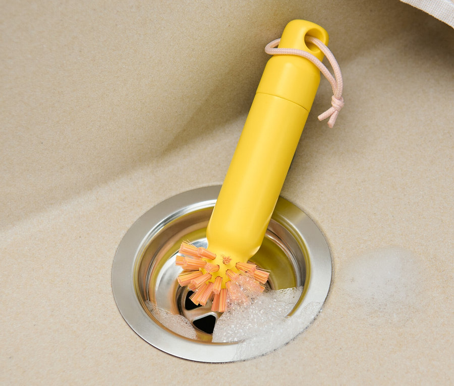 Gelbe 2-in-1-Abflussbürste in einem Edelstahlspülbecken mit Schaum und einem beigen Tuch.