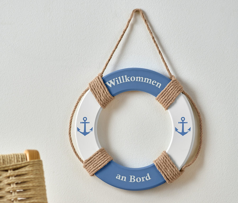 Ein blau-weißer Deko-Rettungsring mit der Aufschrift "Willkommen an Bord" und Anker-Bildern hängt an einer weißen Wand.
