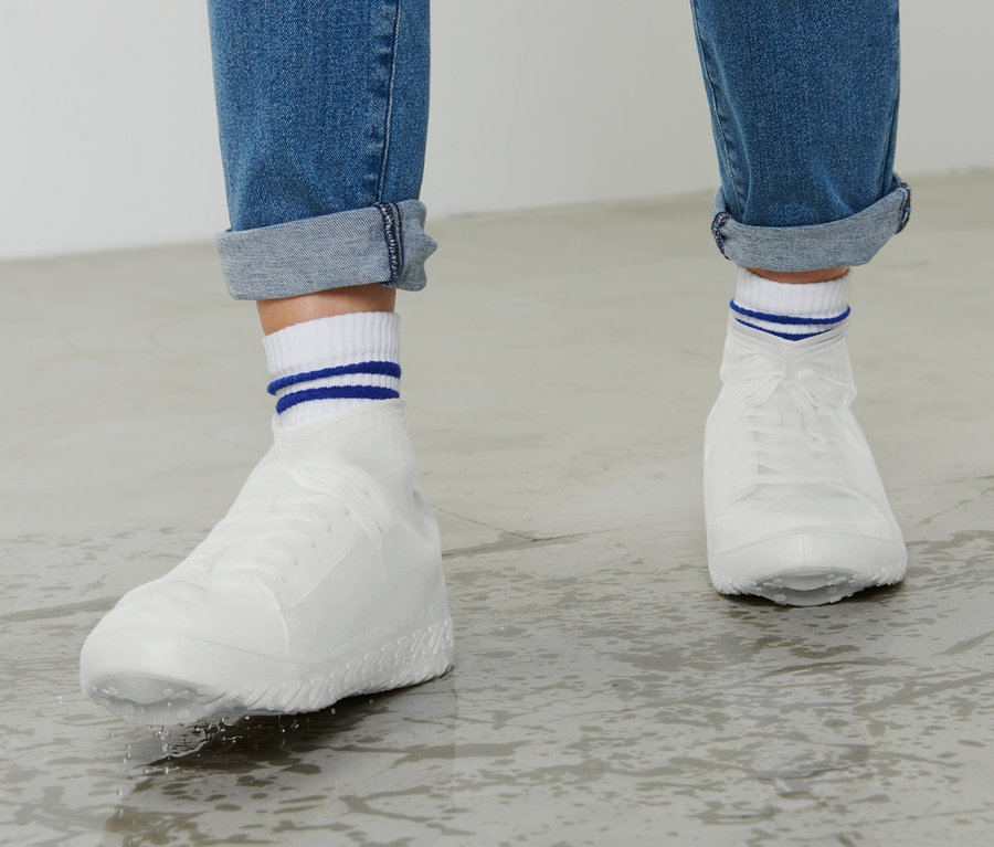Beine in Jeans und weißen Schuhen mit Regen-Überziehschuhe stehen auf nassem Boden.
