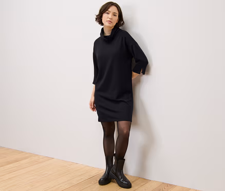 Frau steht an der Wand und trägt ein Sweatkleid aus Modal-Mix, eine schwarze Strumpfhose und schwarze Stiefel.