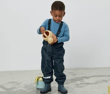 Ein Junge in einer Kinder-Regenhose gießt Wasser mit einer Gießkanne auf den Boden.