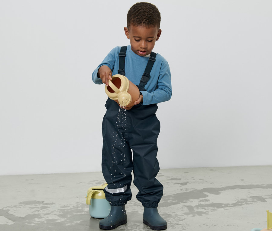 Ein Junge in einer Kinder-Regenhose gießt Wasser mit einer Gießkanne auf den Boden.