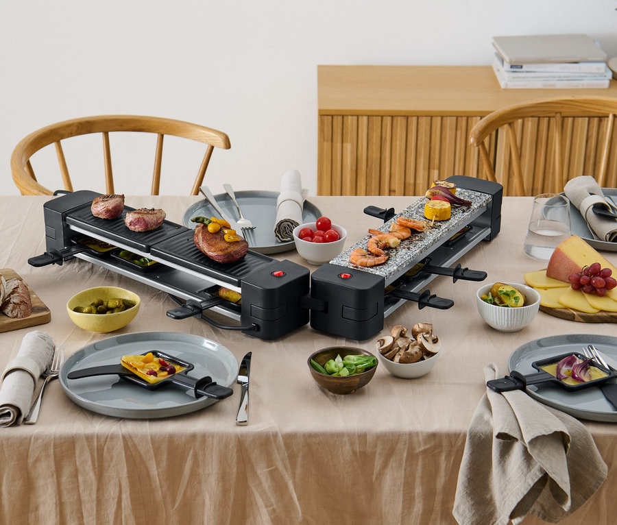 Zwei klappbare Raclette-Grills mit Speisen auf einem Tisch.