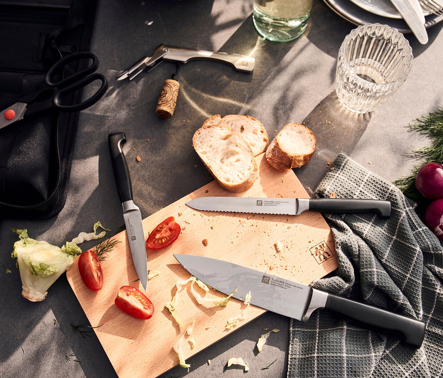Auf einem Tisch liegen eine Schere, ein Weinöffner, ein Korken, Brot, ein Glas, Dill, rote Zwiebeln, ein Geschirrtuch und ein ZWILLING Messer-Traveller-Set.