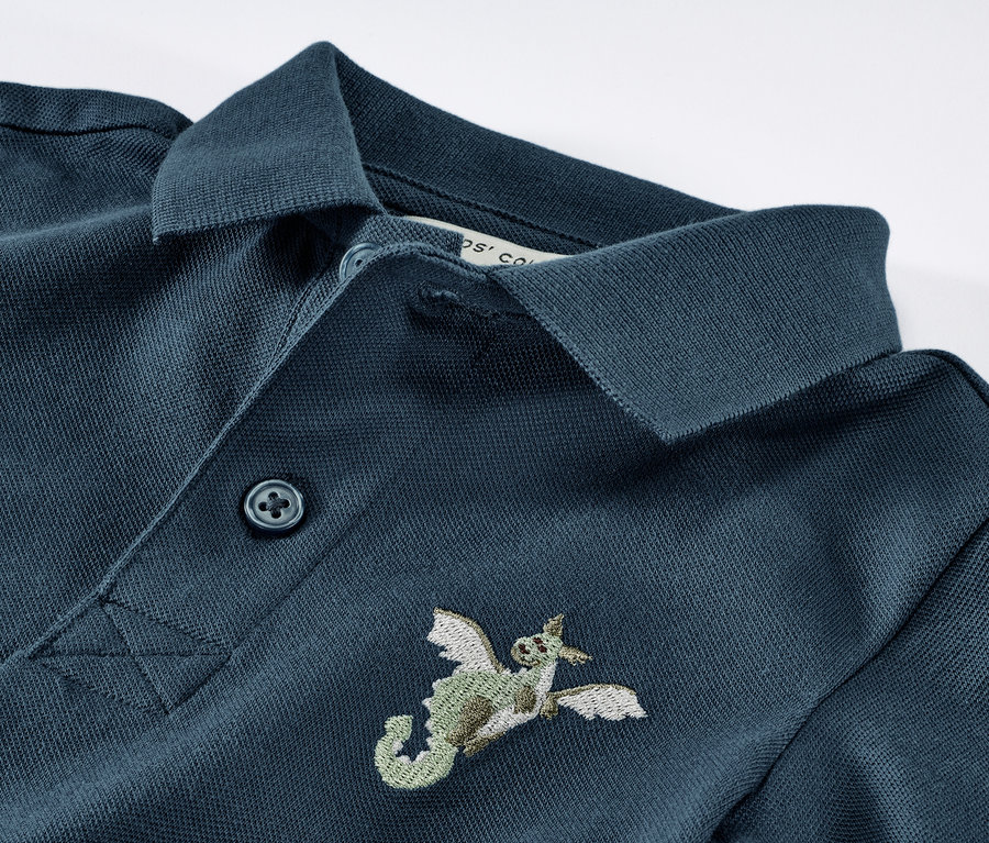 Detailansicht eines blauen Kinder-Poloshirts aus Baumwoll-Piqué mit Drachenstickerei.
