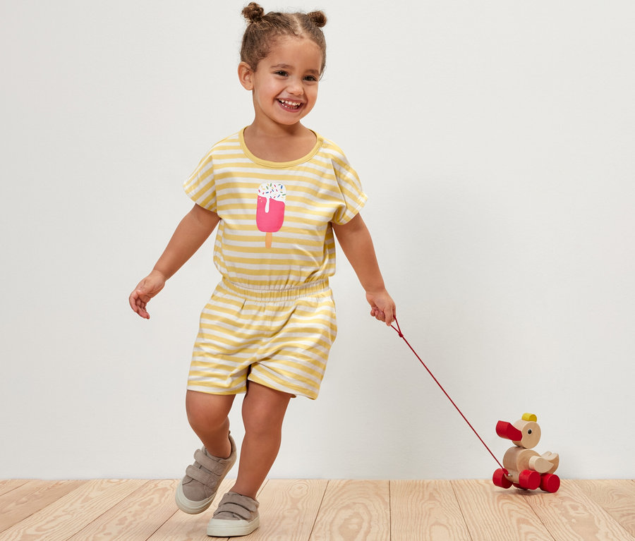 Lächelndes Mädchen im Kinder-Jumpsuit mit Streifen zieht ein Holzspielzeug-Ente hinter sich her.