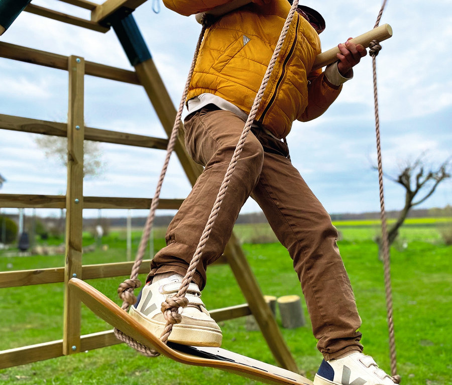 Ein Junge in einer gelben Jacke balanciert auf einer small foot Skateboard-Schaukel auf einem Spielplatz.