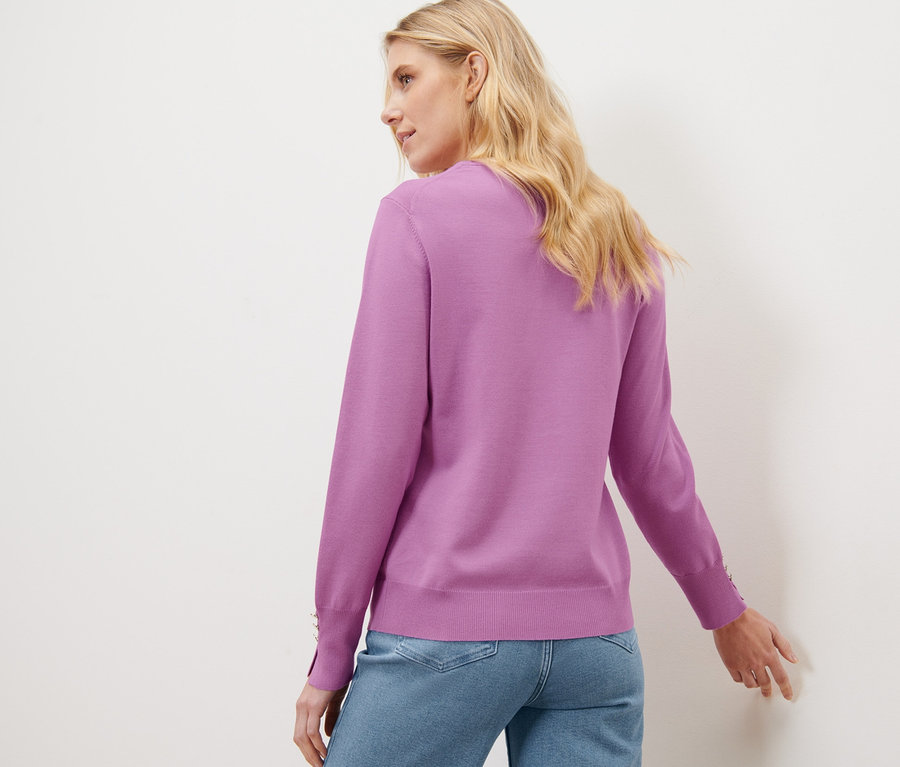 Blonde Frau posiert mit dem Rücken zur Kamera und trägt einen lila Feinstrickpullover und Jeans.