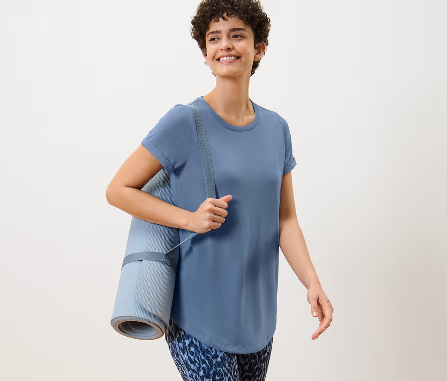 Frau mit braunen Haaren trägt eine Yogamatte und ein Longshirt in Taubenblau.