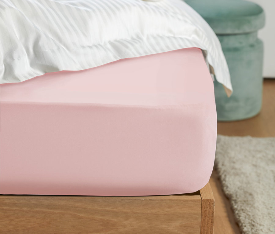 Detailaufnahme eines rosa Spannbetttuchs auf einem Bett mit weiß gestreifter Bettwäsche und einem grünen Hocker im Hintergrund.