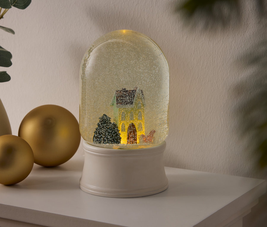 Eine Schneekugel mit Haus und Bäumen auf einem weißen Sockel steht auf einem Regal neben goldenen Kugeln und einer Vase mit Pflanzen.