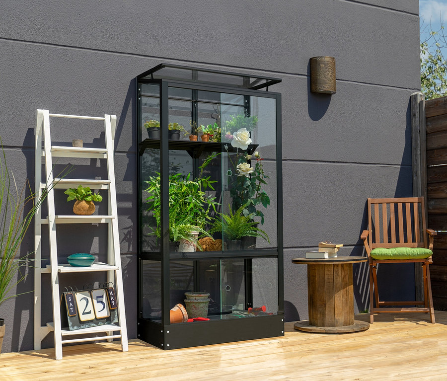 Ein schwarzes Vitavia Balkongewächshaus mit Standfundament steht auf einer Holzterrasse neben einem weißen Regal.