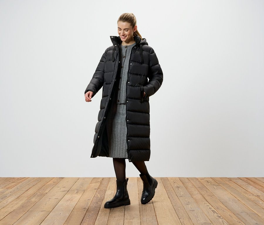 Eine Frau in einer langen, schwarzen Daunenjacke, einem grauen Strickkleid und schwarzen Stiefeln steht auf einem Holzboden.