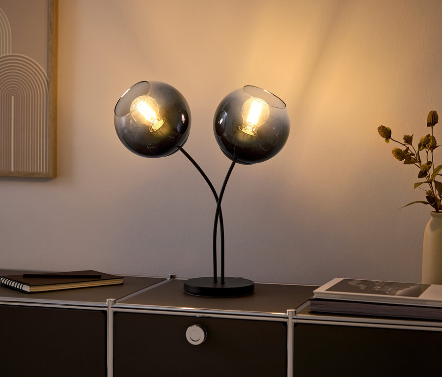 Zwei Glaslampen mit Glühbirnen auf einer schwarzen Kommode.