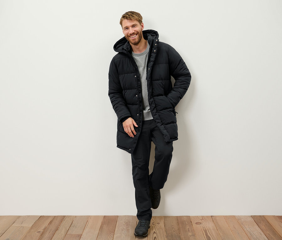 Ein lächelnder Mann mit Bart lehnt an einer Wand und trägt eine schwarze Daunenjacke mit Kapuze, eine schwarze Hose und Stiefel.