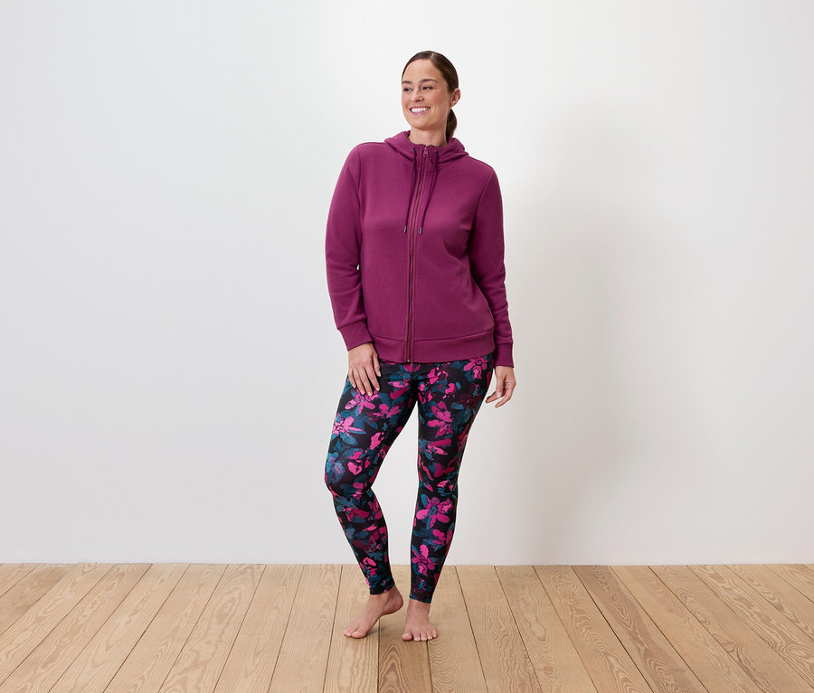 Frau mit bordeauxrotem Reißverschluss-Hoodie und geblümten Leggings steht auf Holzboden.