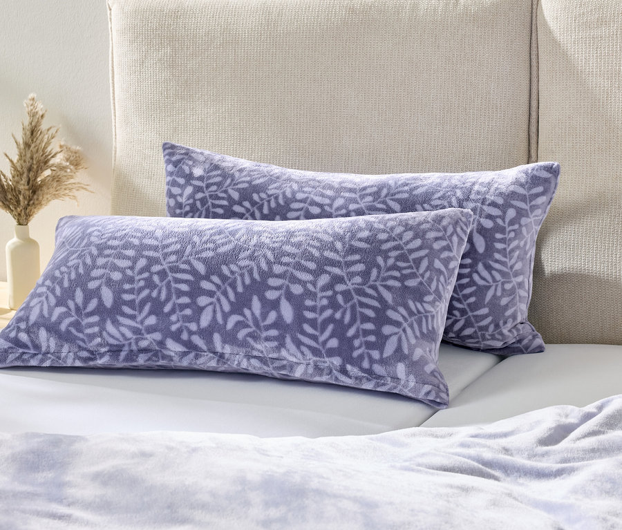 Zwei lila Kissen mit Blättermuster liegen auf einem weißen Bett neben einem Nachttisch mit einer Vase Trockenblumen.