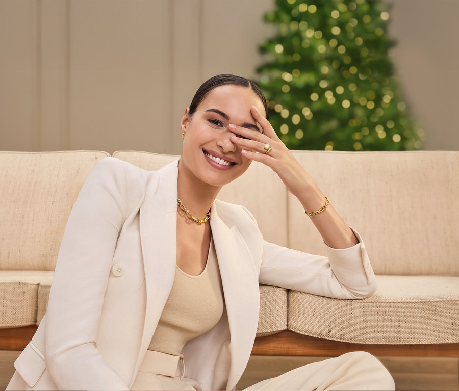 Eine Frau sitzt auf einem Sofa, lächelt und bedeckt ein Auge mit ihrer Hand. Sie trägt ein Jackett, eine Halskette und ein Armband.