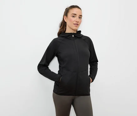 Frau mit Händen in den Taschen einer schwarzen Sportjacke mit Kapuze und Reißverschluss.