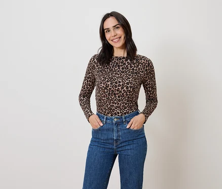 Frau mit braunem Haar posiert in einem Oberteil mit Tiermuster und blauen Jeans.