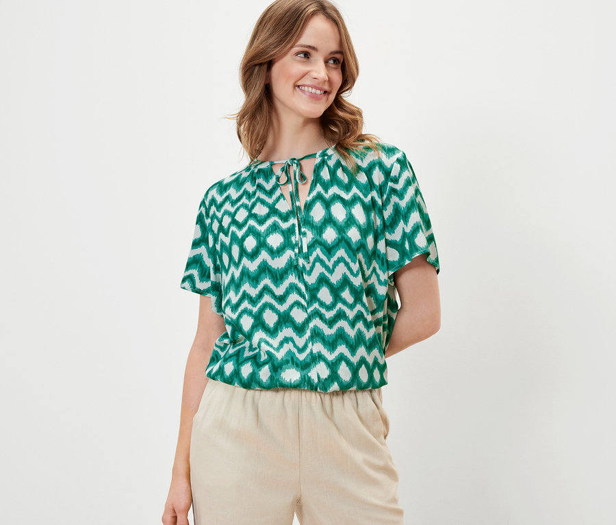 Frau trägt eine bedruckte Tunika-Bluse und eine beige Hose.