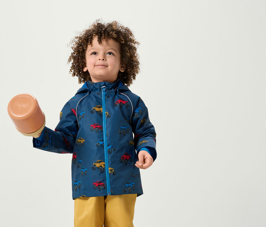 Ein Junge mit lockigem Haar hält einen braunen Behälter. Er trägt eine Jacke mit einem Autoaufdruck und gelbe Hosen.