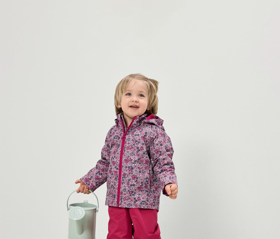 Kleinkind hält eine kleine grüne Gießkanne. Es trägt eine Jacke mit Blumenmuster und eine pinke Hose.