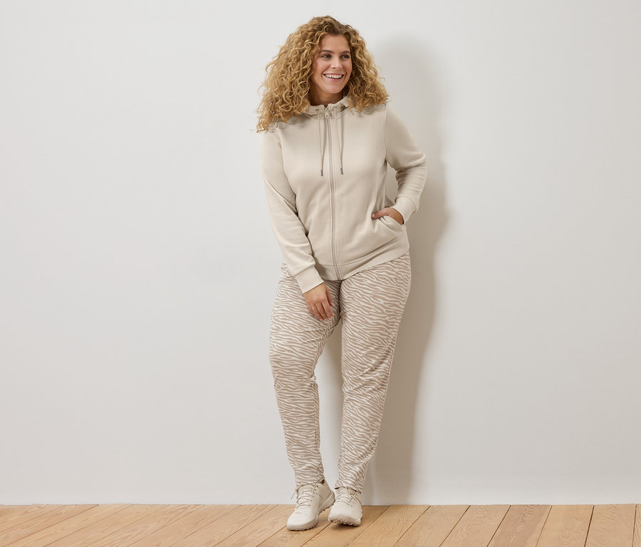 Frau mit lockigem Haar trägt eine beige Kapuzenjacke und eine Hose mit Zebra-Print und lehnt an einer weißen Wand.
