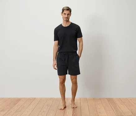 Mann steht barfuß vor einer weißen Wand und trägt ein schwarzes T-Shirt und Shorts.