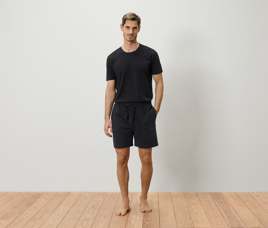 Mann steht barfuß vor einer weißen Wand und trägt ein schwarzes T-Shirt und Shorts.